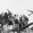 A group of German children stand atop building rubble, cheering a United States cargo airplane